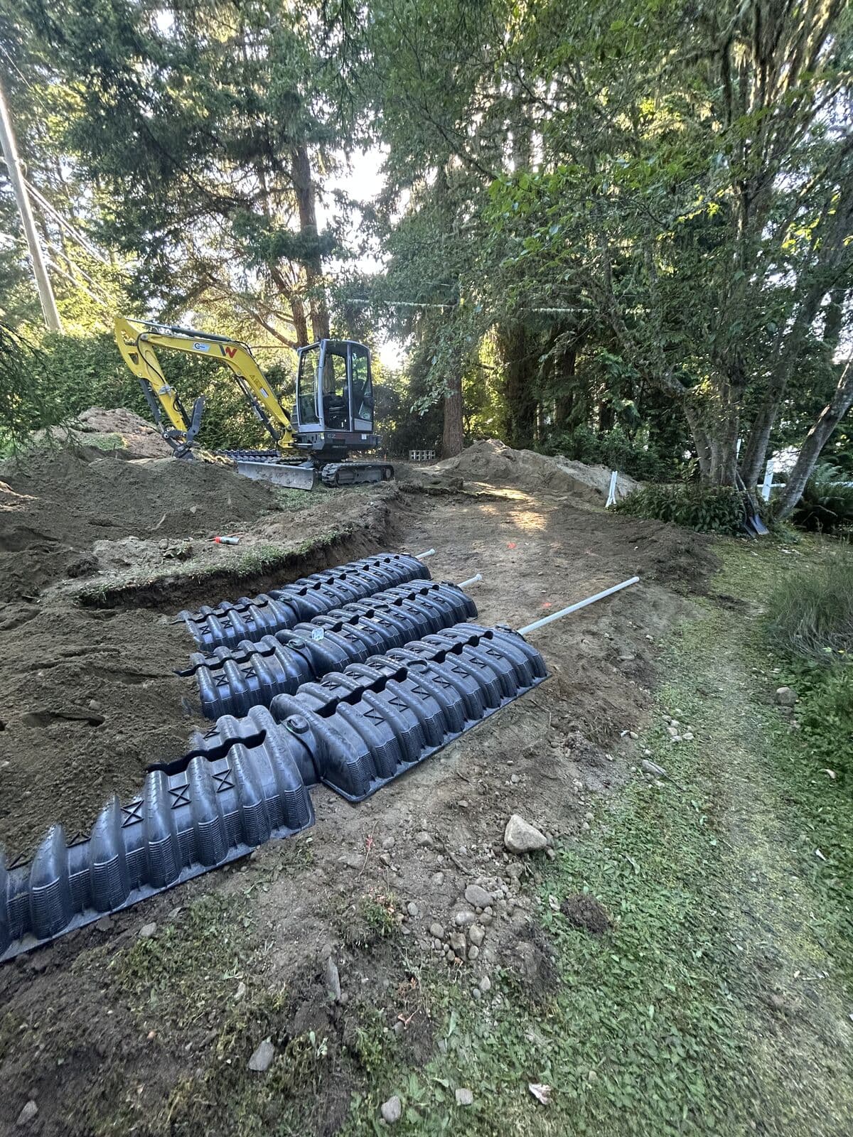 Three parallel rows of black plastic infiltration chambers laid end to end on excavated ground, with white PVC pipes running across them. A yellow excavator sits on the spoil bank in the background among mature trees.
