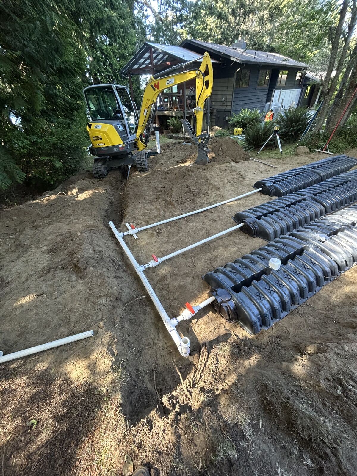 A white PVC distribution manifold with several red-handled ball valves laid in a shallow trench, with branch lines running to three rows of black infiltration chambers on the right. A yellow excavator and a house with a covered deck are in the background.