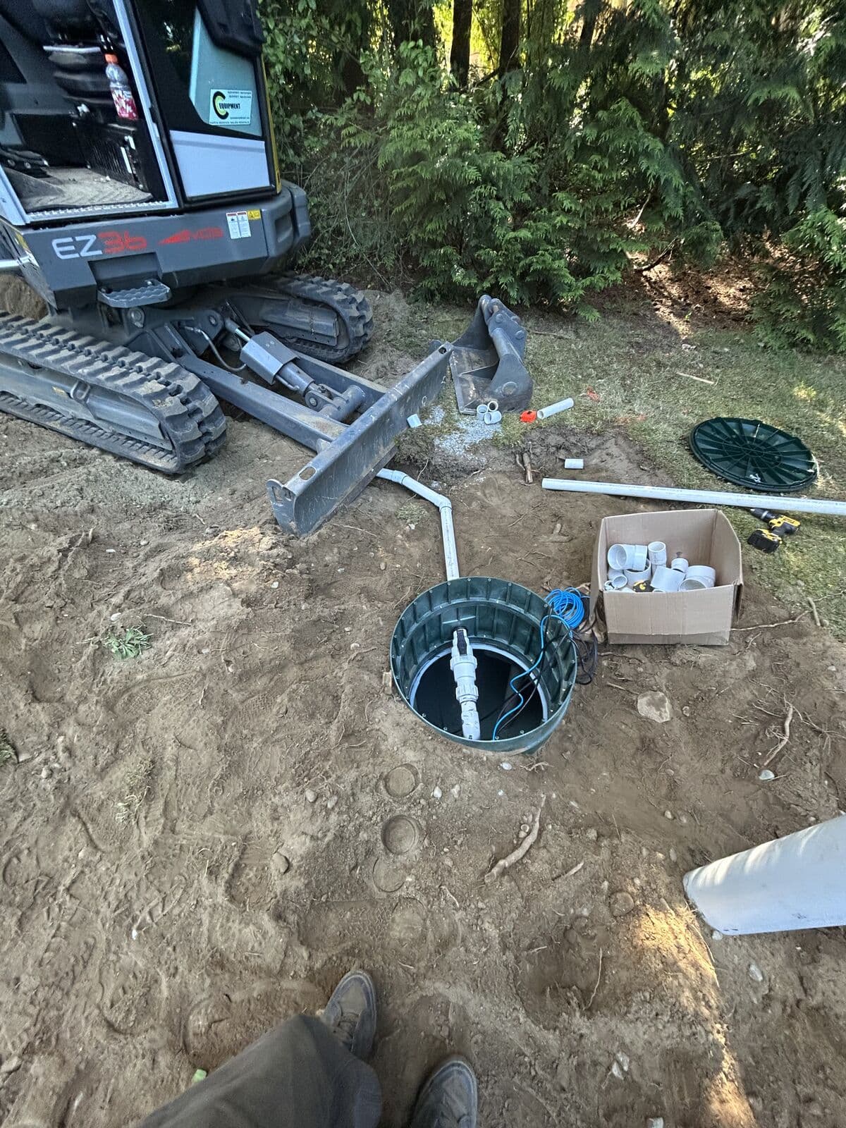 An open green cylindrical pump chamber riser set into bare ground, containing a vertical white PVC discharge pipe with a union fitting and blue and black electrical float leads. Its lid lies on the grass nearby beside a box of PVC fittings, with an excavator parked at the left.