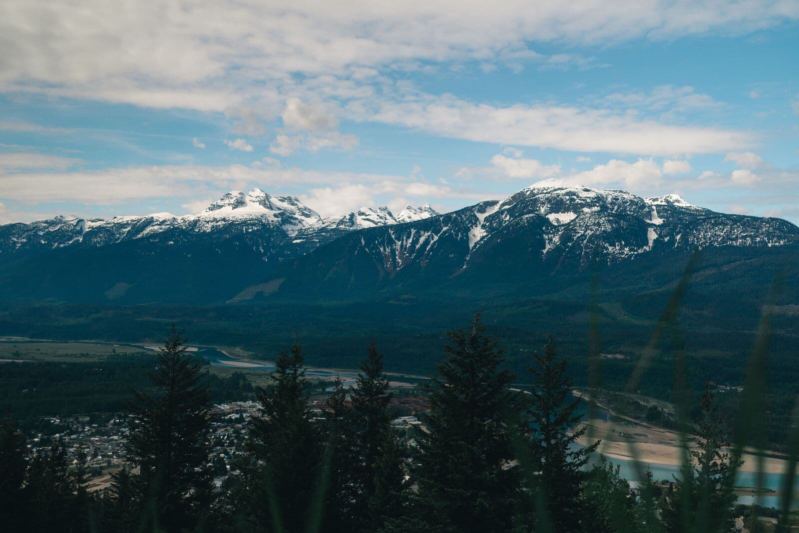 Snow-capped mountains under a broken cloudy sky, seen across a wide valley floor with a river winding through it and conifers in the foreground.