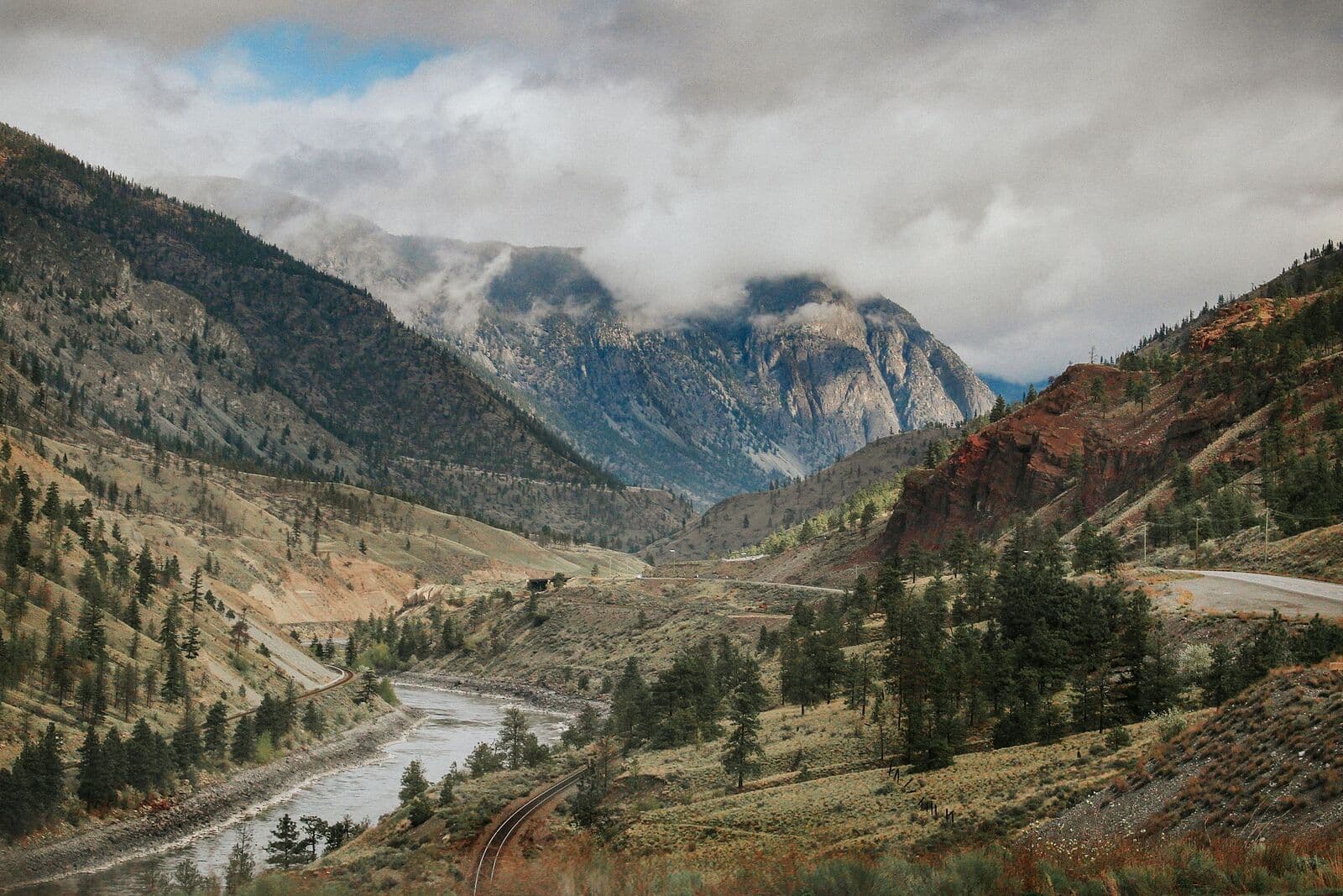 A dry canyon of sparse pines and pale grassland, with a river and a railway line running along the valley floor and red-brown rock bluffs rising to cloud-covered mountains.