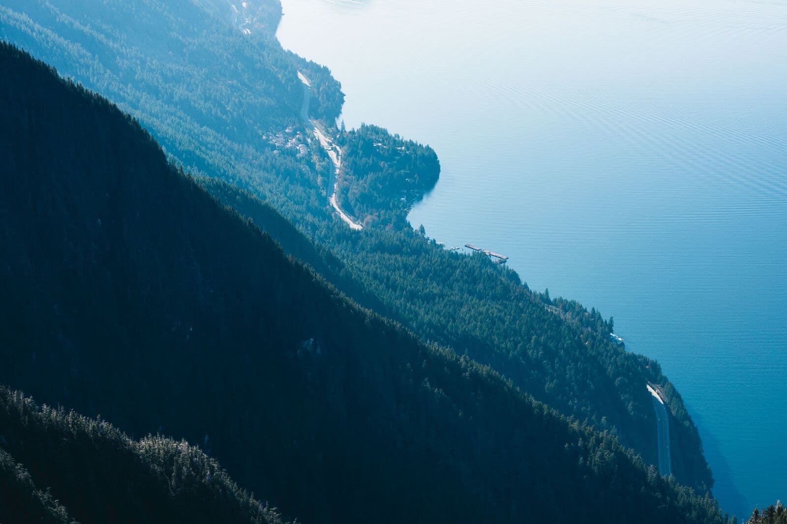 A steep forested mountainside falling away to a deep blue coastal inlet, with a winding shoreline below and forested ridges fading into haze beyond.