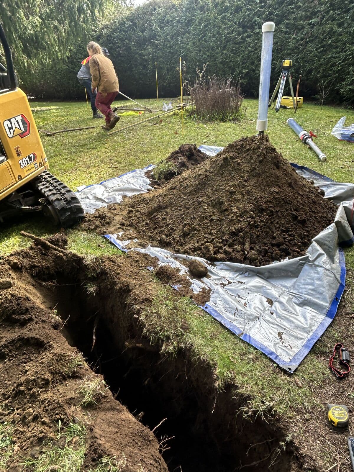 A narrow trench cut through lawn with excavated soil heaped on a blue and silver tarpaulin. A compact tracked excavator stands at the left, a tripod-mounted rotating laser level at the right, and two people work beside marker stakes in front of a tall hedge.