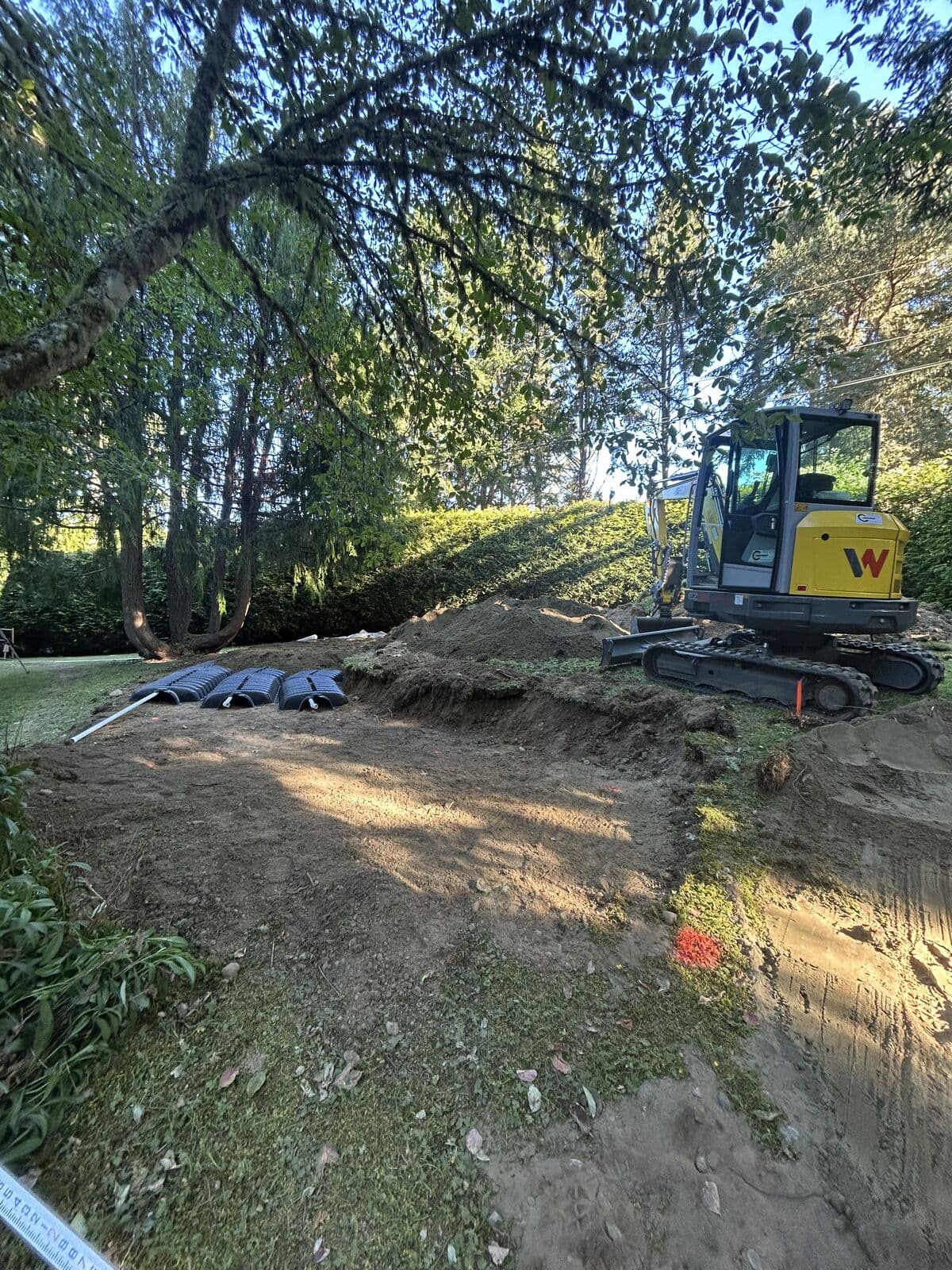 A cleared and levelled area of ground beneath tall conifers, with three black plastic infiltration chambers laid out at the left and loose spoil banked along the right. A yellow tracked excavator sits at the top right beside a clipped hedge.