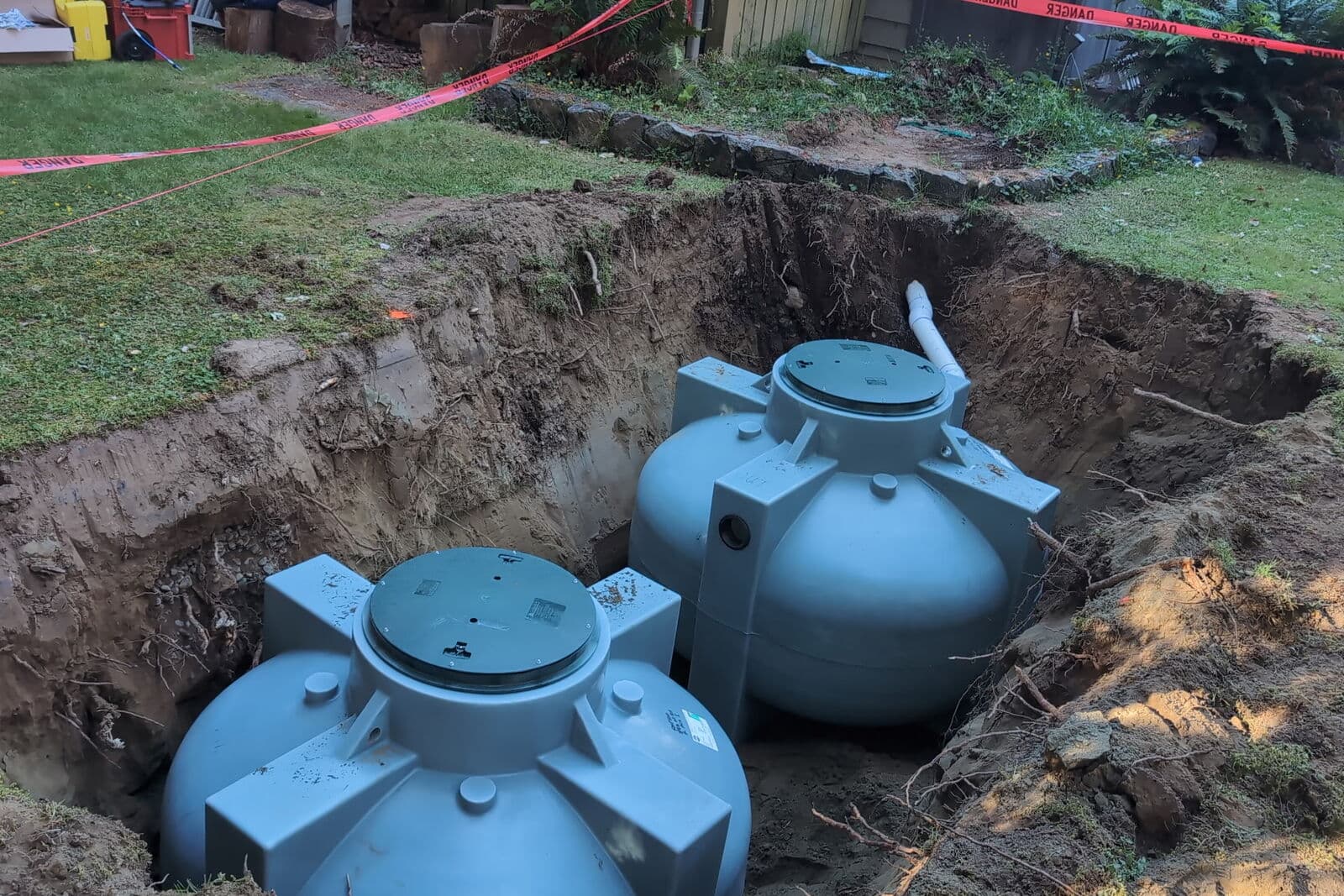 Two pale blue-grey moulded tanks seated in a rectangular excavation cut into a lawn, with a stone retaining wall and red danger tape strung behind them. A white pipe runs into the rear tank, and exposed roots and loose soil line the cut faces of the trench.
